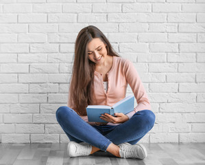 Beautiful young woman with book sitting on floor near brick wall