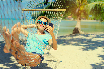 Vacation concept. Enjoying the summer. Young pretty woman in hat and sunglasses using smartphone laying in hammock on the beach.