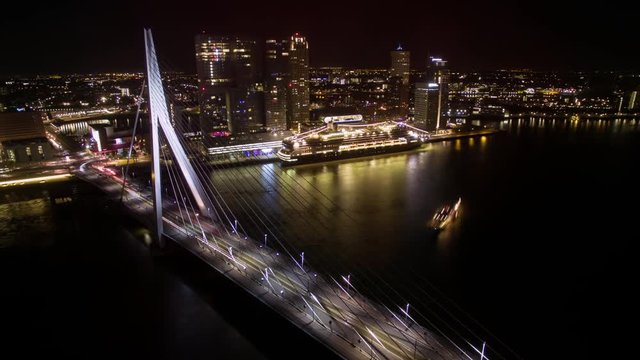 Timelapse shot of transport traffic in Erasmus Bridge and ship sailing along the river on background of illuminated buildings of Rotterdam at night