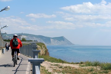 Fototapeta na wymiar Cyclist in red sweater, protective helmet and head athletic backpack riding bicycle along seaside promenade of resort town Anapa on sunny spring day. Over mountains and sea - haze