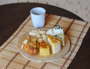 Cheese plate and a cup of coffee. Assorted pickled cheeses on round wooden board on the mat