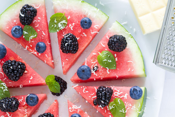 Sliced watermelon pizza with blueberries, blackberries , mint leaf and white chocolate crisps, above view.