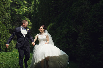 Bride raises her dress while she runs with groom through the green garden