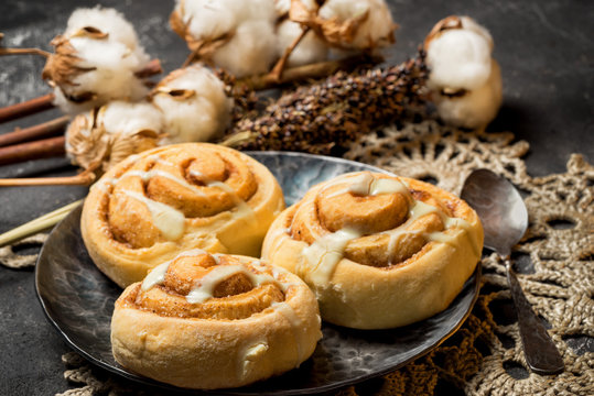 three icing cinnamon rolls on a metal plate with spoon and crocheted doily on a dark cement background with cotton boxes behind.