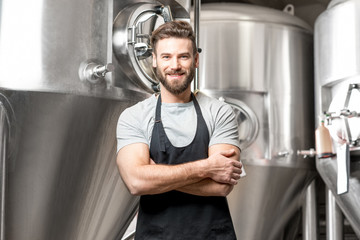 A portrait of handsome brewer in uniform at the beer manufacture with metal containers on the background