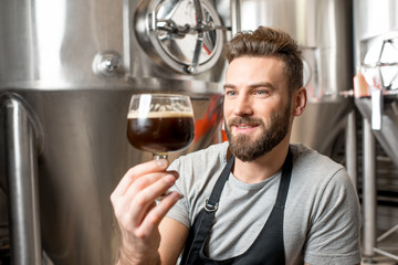 Handsome brewer in uniform checking quality of the beer sitting in the manufacturing with metal containers on the background