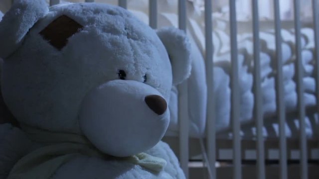 closeup big white toy bear sits near white empty baby crib on background at night