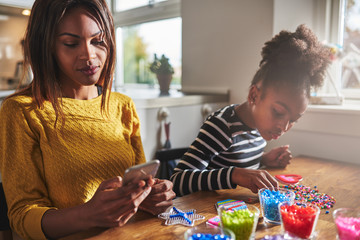 Woman checking phone while child does beading