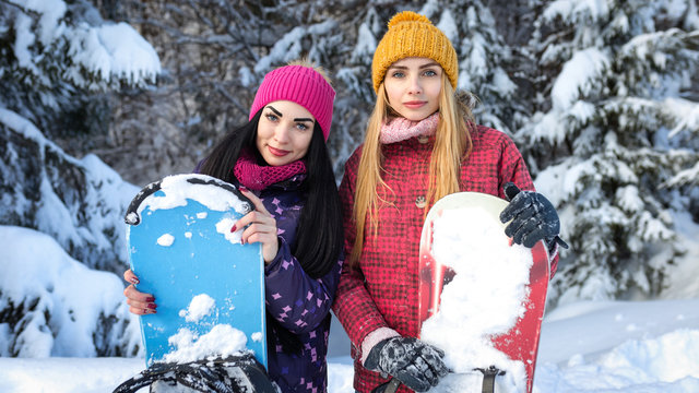 Two attractive girls snowboarders are among snowy fir trees in the winter and keep snowboards hands