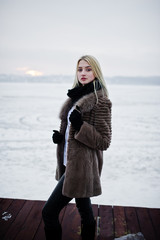 Portrait of young elegance blonde girl in a fur coat at pier bac