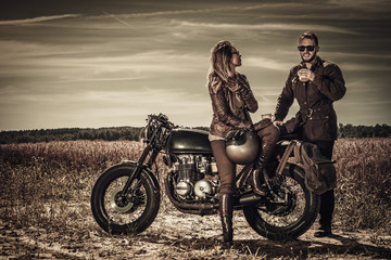 Young, stylish cafe racer couple on vintage custom motorcycles in field