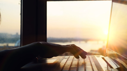 Close up of woman's hands typing on laptop's keyboard during sunset with beautiful sun lense flare effects.