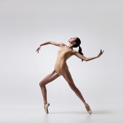 young beautiful ballet dancer in beige swimsuit posing on pointes on light grey studio background