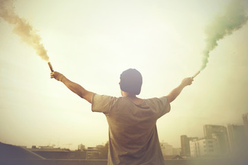 Male teenager holding colorful smoke sticks up in the air over urban city background
