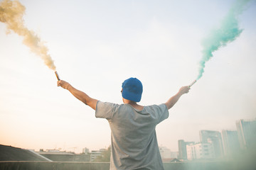 Male teenager holding colorful smoke sticks up in the air over urban city background