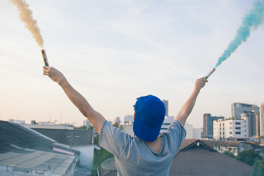 Male teenager holding colorful smoke sticks up in the air over urban city background