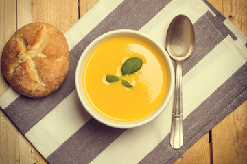 Sopa crema de calabaza zapallo junto a una cuchara y un pan casero sobre un mantel a rayas y una mesa de madera. Vista superior