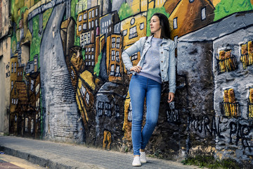 Young and attractive girl is standing next to the graffiti wall and looking somewhere out the camera, Barcelona, Spain