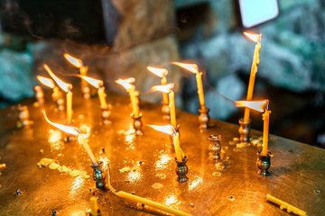 Small wax candles stuck in a tray burn outdoors at blowing wind. Orthodox Christianity religious tradition to set flaming candles while worshiping God. Close up view
