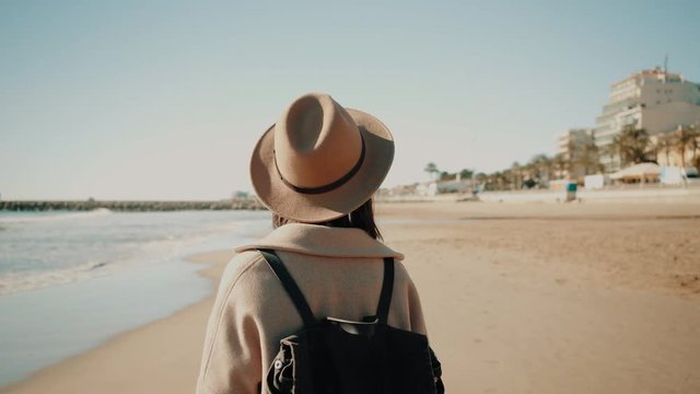 Camera follows attractive smiling girl weared fedora hat and wool coat, walks with black backpack on empty sand beach on shoreline at very sunny day Overexposed high key image