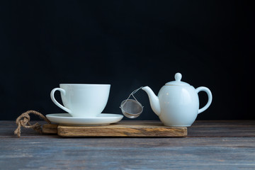 White porcelain tea cup and saucer, teapot and strainer on wooden desk