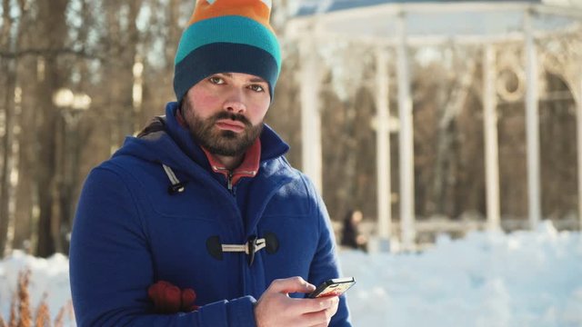 young man in blue coat and colorful hat with beard typing text in smartphone, look up and stare to camera without smile, with steam from the mouth because of cold winter day under the sunlight