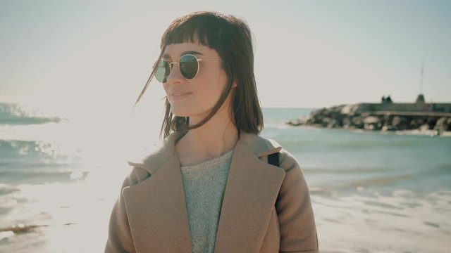 Camera slowly moves back from close up of beautiful smiling brunette girl in wool coat with fedora hat in her hand, smiling at cameta and posing in front of sea on sand beach at sunny day