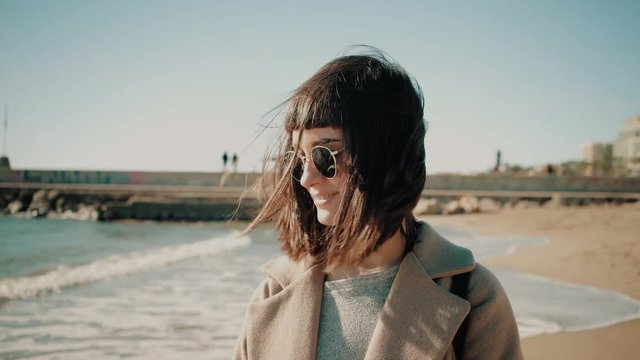 Portrait of sexy and beautiful brunette girl in sunglasses looks at camera, smiles and plays with her dark hairs at sunny windy winter day on empty beach next to sea Escaping from town to quiet place