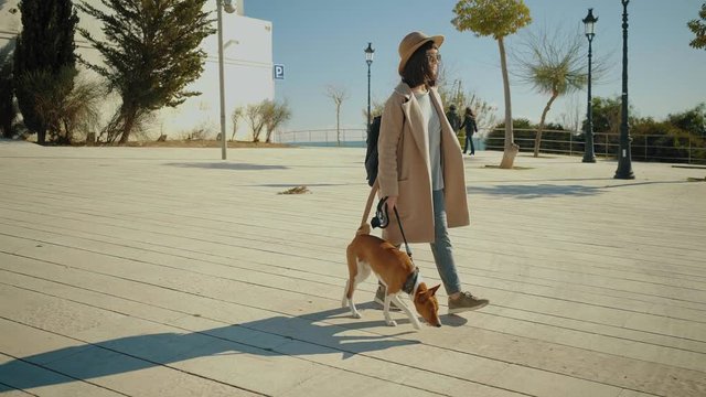 Attractive brunette girl walks with her beautiful and curious basenji dog on leash, at windy and sunny day in small town near sea