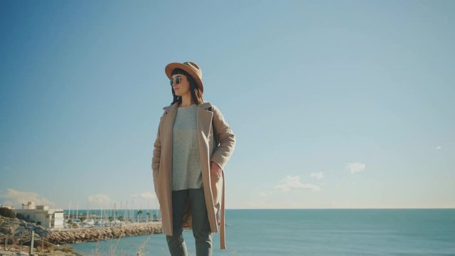 Beautiful brunette girl stays on stone railing and takes photos with her smartphone at bright sunny day next to the sea, overexposed style image