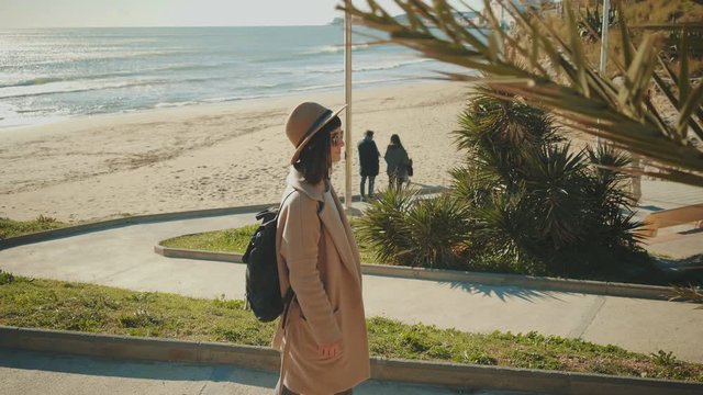 Camera follows by side attractive brunette girl in sunglasses and with black backpack on her back walking down to sand beach next to sea through small narrow lane with green palms at sunny day.