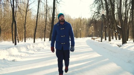 young man in blue coat and colorful hat walk in snowy winter park, look at his smartphone, see horrify message, shout at phone and run away very fast, camera follows after him