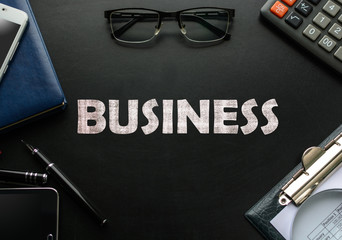 Black chalkboard with business accessories (notepad, magnifying glass, fountain pen, tablet, phone, glasses and calculator) and text BUSINESS. Top view.