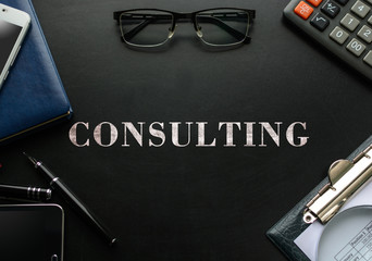 Black chalkboard with business accessories (notepad, magnifying glass, fountain pen, tablet, phone, glasses and calculator) and text CONSULTING. Top view.