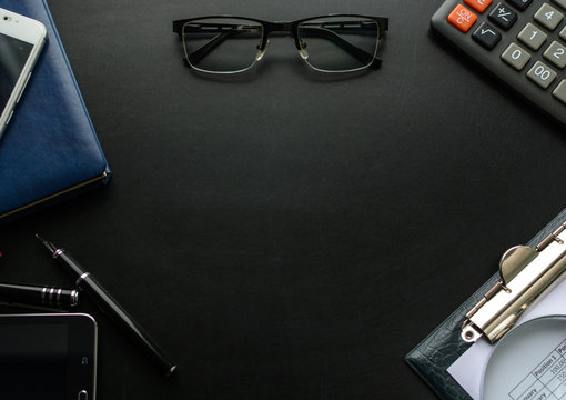 Black chalkboard with business accessories (notepad, magnifying glass, fountain pen, tablet, phone, glasses and calculator) and free space for your text. Top view.