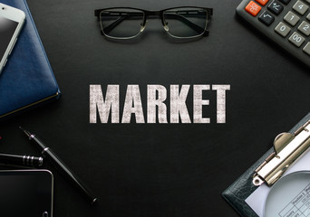 Black chalkboard with business accessories (notepad, magnifying glass, fountain pen, tablet, phone, glasses and calculator) and text MARKET. Top view.