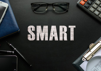 Black chalkboard with business accessories (notepad, magnifying glass, fountain pen, tablet, phone, glasses and calculator) and text SMART. Top view.