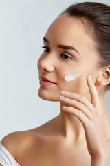 Naturally beautiful brunette woman with flawless skin applying care cream to her face over gray background