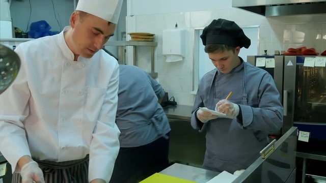 Young trainee watching how chef is cooking and taking notes