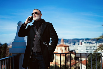 Serious attractive man wearing black jacket and talking at mobile phone while spending time in city park.Horizontal,blurred background.