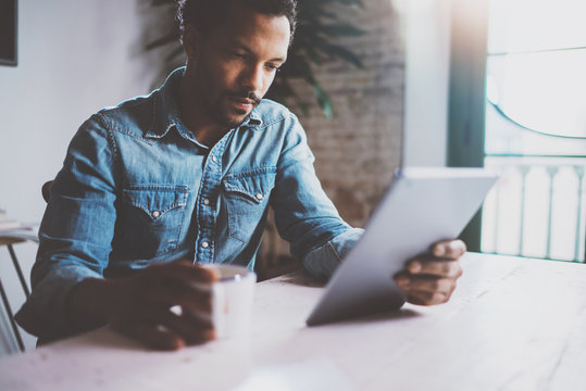 Serious young African man reading world news by digital tablet while sitting at the table on a sunny morning.Concept of coworking people working home.Blurred background,flare effect.