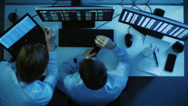 Top View of Two Stock Brokers Looking and Analysing Market Numbers. Before Them Two Monitors with Stock Exchange Numbers, Graphs. Shot on RED EPIC-W 8K Helium Cinema Camera.