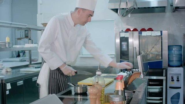 Chef slicing garlic on the cutting board with a knife and mixing it with salt brought by another cook