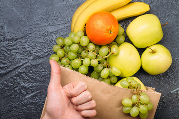 Food background of fresh fruit bananas oranges apples and grapes in shopping bag and man hand with thumb up on black table Healthy lifestyle Top view Copy space