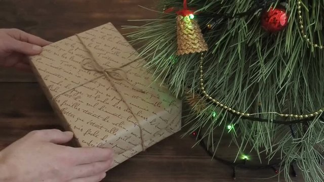 The man puts gift boxes under the Christmas tree. Fir stand on the wooden table, decorated glowing garland and Christmas toys. Boxes with gift wrapped in brown and colored paper and tied with twine.