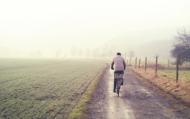Obraz premium Old man on a bicycle, winter scene with dirt road, fields and fog. Tranquil scene.