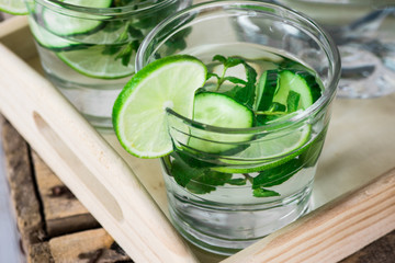 Glasses with detox infused cucumber water with lime and mint on wooden tray, pitcher,close up