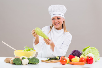 Portrait of cheerful female chef sitting at the table and showing zucchini while making healthy meal.
