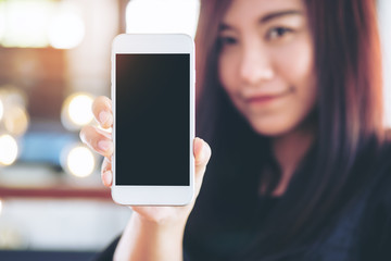 Mockup image of a beautiful woman holding and showing white mobile phone with blank black screen and smiley face in vintage cafe