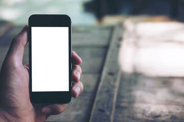 Mockup image of hands holding black mobile phone with blank white screen and vintage wood table background